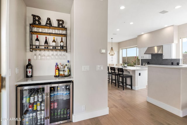 bar with backsplash, light wood-style floors, beverage cooler, a bar, and wall chimney exhaust hood