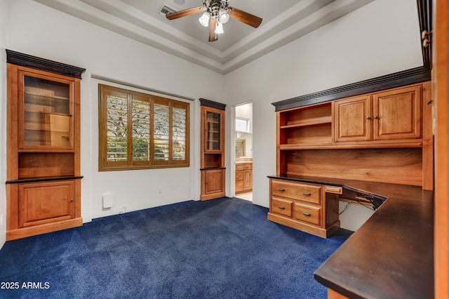 unfurnished office featuring a raised ceiling, ceiling fan, built in desk, and dark carpet