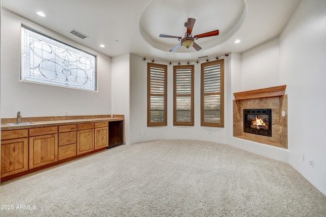 unfurnished living room with sink, light carpet, a raised ceiling, ceiling fan, and a tiled fireplace