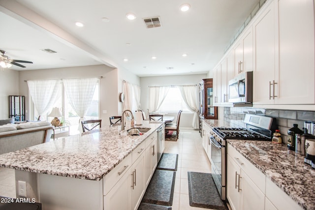 kitchen with appliances with stainless steel finishes, an island with sink, backsplash, white cabinets, and light stone counters