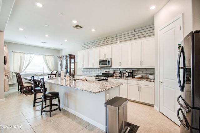 kitchen with light stone countertops, appliances with stainless steel finishes, white cabinets, decorative backsplash, and a kitchen island with sink