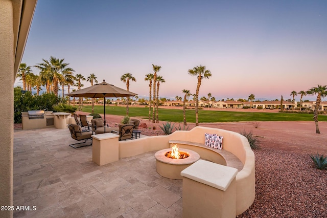 patio terrace at dusk featuring exterior kitchen, a lawn, grilling area, and a fire pit