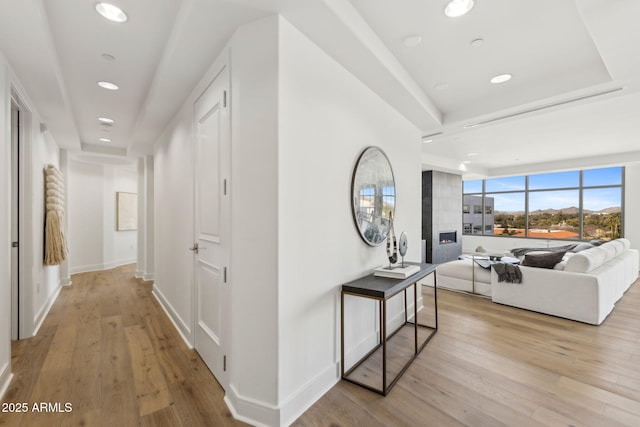 hall featuring baseboards, light wood finished floors, a raised ceiling, and recessed lighting