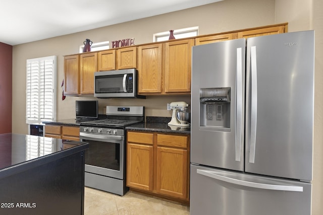 kitchen with appliances with stainless steel finishes, brown cabinetry, dark stone countertops, and light tile patterned floors