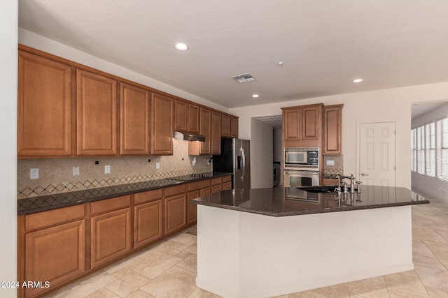 kitchen featuring sink, decorative backsplash, dark stone countertops, an island with sink, and appliances with stainless steel finishes