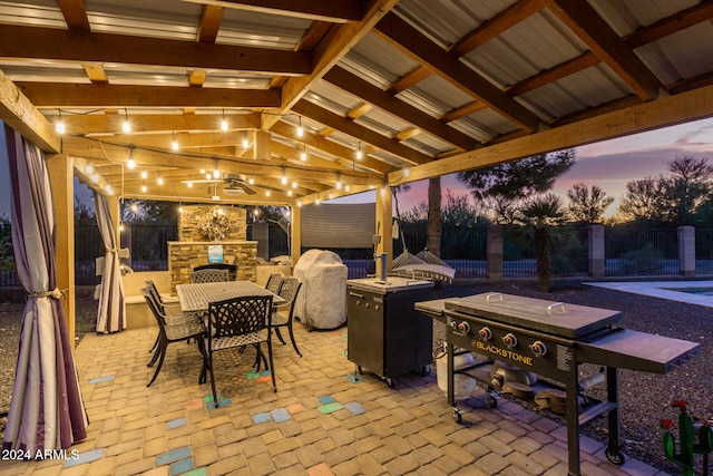 patio terrace at dusk with ceiling fan, an outdoor stone fireplace, and an outdoor kitchen