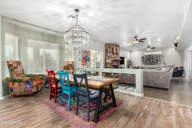 dining space with a stone fireplace, wood-type flooring, and ceiling fan with notable chandelier