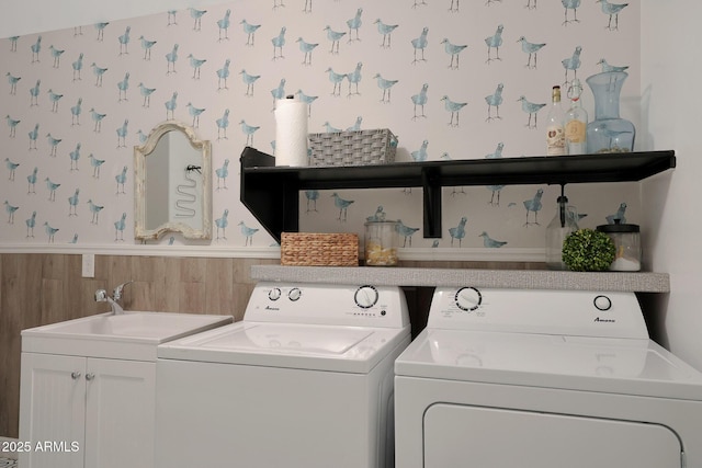 clothes washing area with wallpapered walls, a wainscoted wall, washing machine and dryer, cabinet space, and a sink