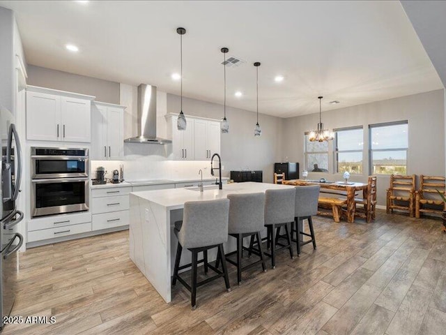 kitchen featuring pendant lighting, wall chimney range hood, sink, white cabinets, and a center island with sink