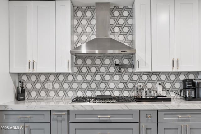 kitchen with wall chimney range hood, gray cabinets, light stone countertops, stainless steel gas cooktop, and tasteful backsplash