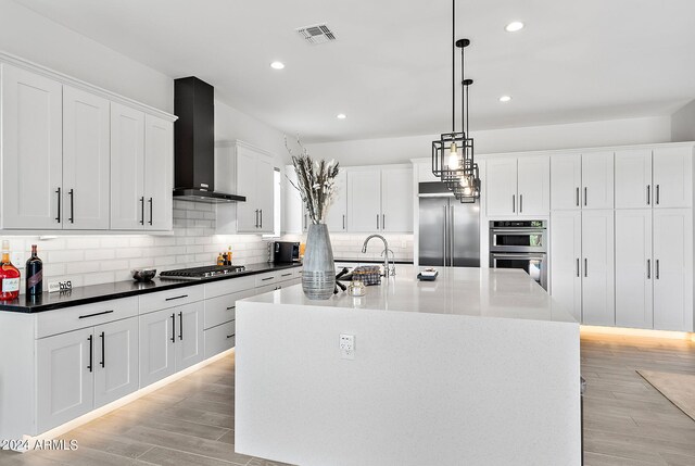 kitchen with appliances with stainless steel finishes, light wood-type flooring, wall chimney range hood, an island with sink, and white cabinetry