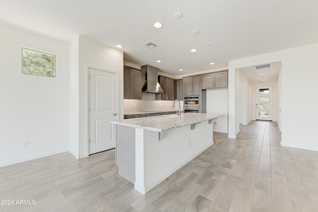 kitchen with light stone counters, a center island with sink, wall chimney exhaust hood, stainless steel double oven, and light wood-type flooring