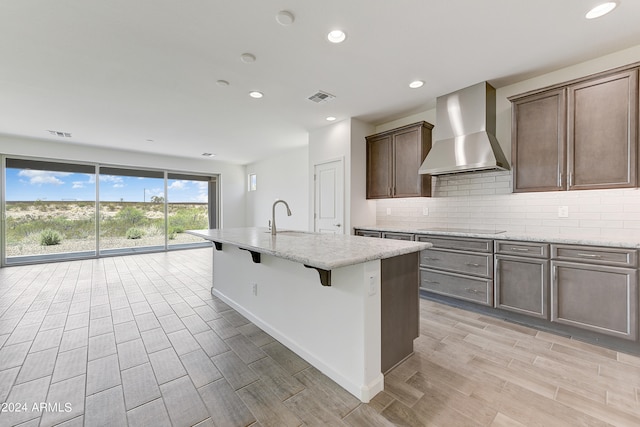 kitchen with black electric cooktop, wall chimney range hood, light hardwood / wood-style floors, and an island with sink