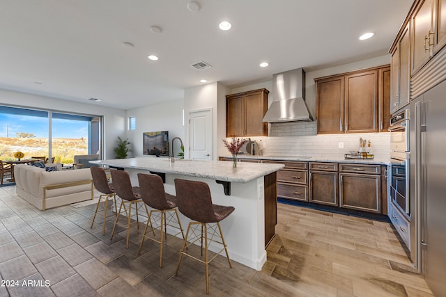 kitchen featuring an island with sink, light wood-type flooring, light stone counters, wall chimney range hood, and a breakfast bar area