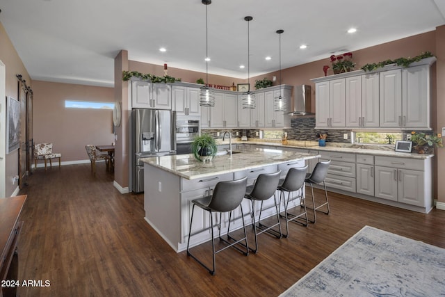 kitchen featuring a center island with sink, stainless steel refrigerator with ice dispenser, hanging light fixtures, wall chimney exhaust hood, and white cabinetry