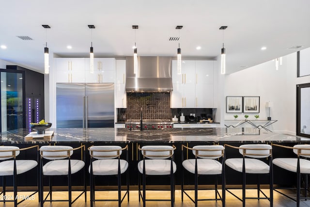 kitchen featuring wall chimney range hood, a kitchen bar, high end fridge, white cabinets, and dark stone countertops