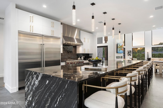 kitchen featuring decorative backsplash, wall chimney range hood, dark stone counters, pendant lighting, and appliances with stainless steel finishes