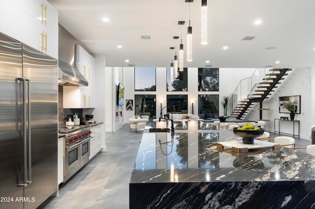 kitchen featuring wall chimney range hood, high quality appliances, hanging light fixtures, dark stone counters, and white cabinets