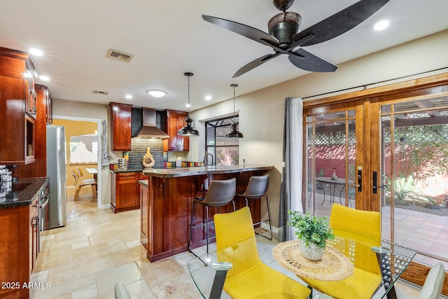 kitchen with a breakfast bar area, decorative light fixtures, stainless steel fridge, decorative backsplash, and wall chimney range hood