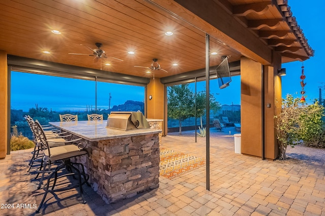 patio terrace at dusk with grilling area, an outdoor kitchen, outdoor wet bar, and a ceiling fan