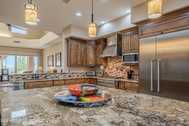 kitchen with built in appliances, light stone counters, a sink, wall chimney range hood, and pendant lighting