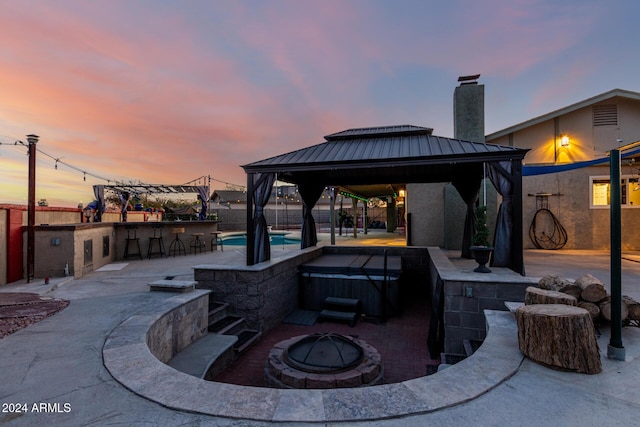 patio terrace at dusk featuring a gazebo, an outdoor bar, a hot tub, and an outdoor fire pit