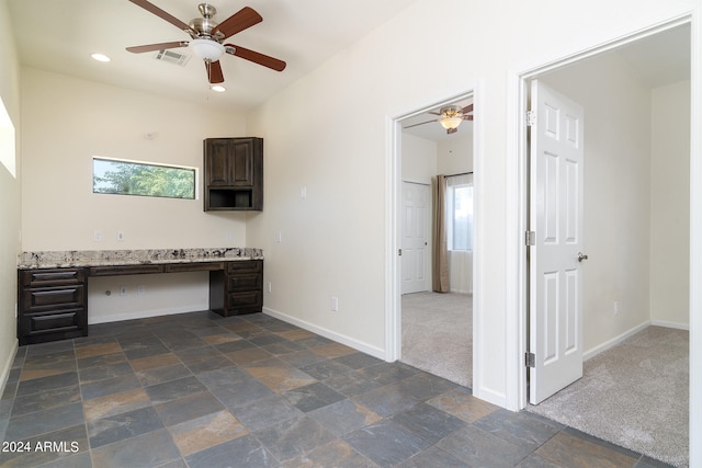 unfurnished office with built in desk, ceiling fan, dark colored carpet, and a wealth of natural light