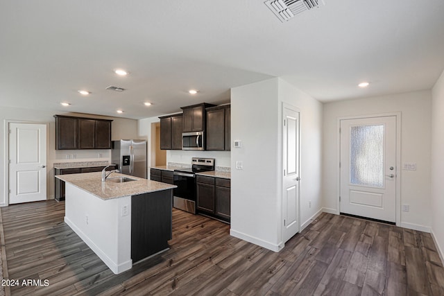 kitchen with light stone counters, dark hardwood / wood-style floors, stainless steel appliances, a center island with sink, and dark brown cabinets