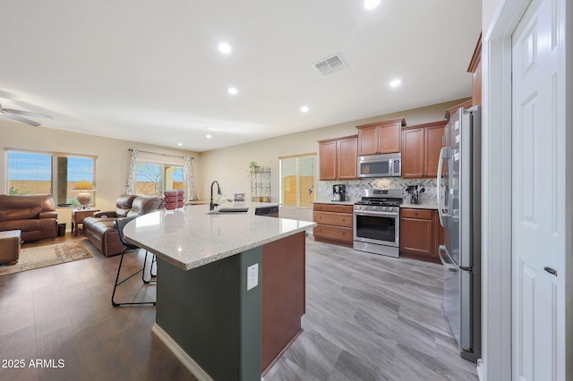 kitchen with a breakfast bar, a center island with sink, appliances with stainless steel finishes, light stone countertops, and backsplash