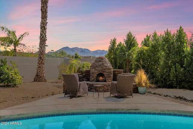 pool at dusk featuring a mountain view, a patio area, and an outdoor stone fireplace