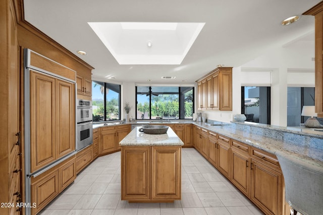 kitchen with kitchen peninsula, stainless steel double oven, a kitchen island, a raised ceiling, and light stone countertops