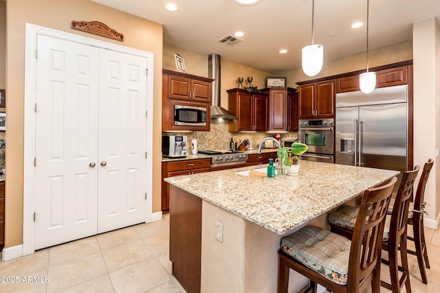 kitchen with sink, built in appliances, a center island with sink, decorative light fixtures, and wall chimney exhaust hood