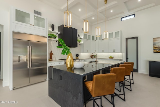 kitchen with stainless steel built in refrigerator, a kitchen island with sink, a high ceiling, coffered ceiling, and white cabinetry