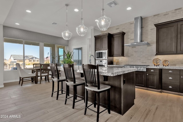 kitchen with wall chimney exhaust hood, decorative light fixtures, stainless steel appliances, light wood-type flooring, and a center island with sink