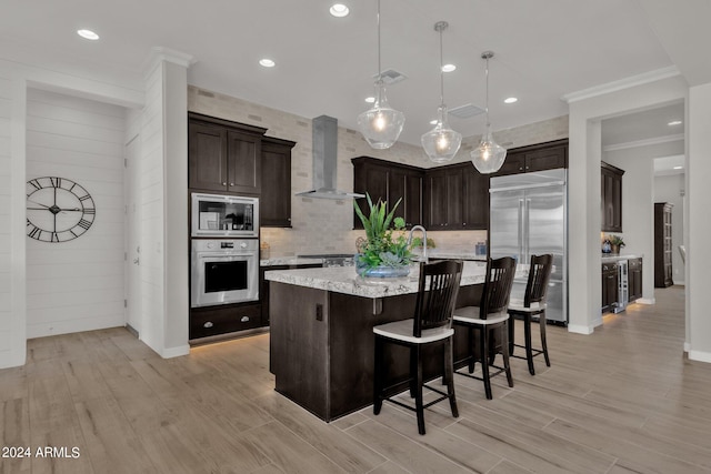 kitchen featuring an island with sink, dark brown cabinets, hanging light fixtures, built in appliances, and wall chimney exhaust hood