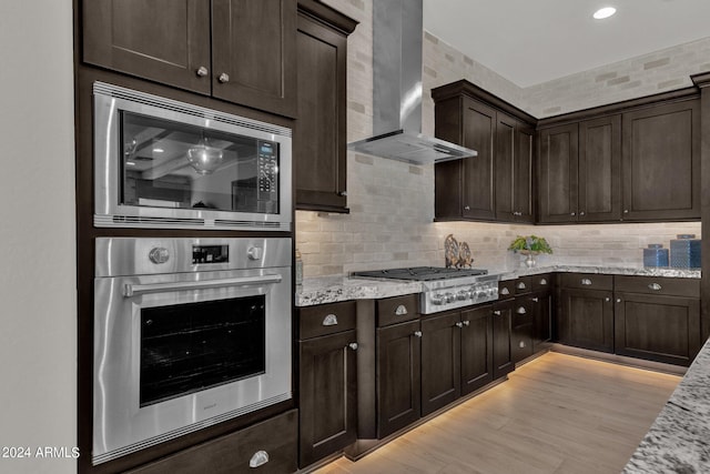 kitchen with appliances with stainless steel finishes, decorative backsplash, light wood-type flooring, wall chimney range hood, and light stone counters