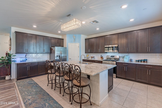 kitchen with dark brown cabinets, a kitchen island with sink, light stone counters, and stainless steel appliances