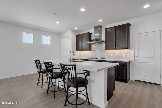 kitchen with stainless steel gas stovetop, a kitchen island with sink, light hardwood / wood-style floors, wall chimney exhaust hood, and sink