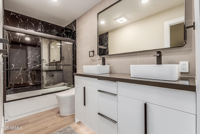 full bathroom with combined bath / shower with glass door, tile walls, vanity, wood-type flooring, and toilet