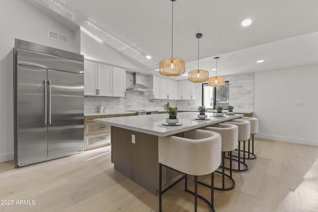 kitchen with wall chimney exhaust hood, white cabinetry, decorative light fixtures, built in fridge, and a kitchen island