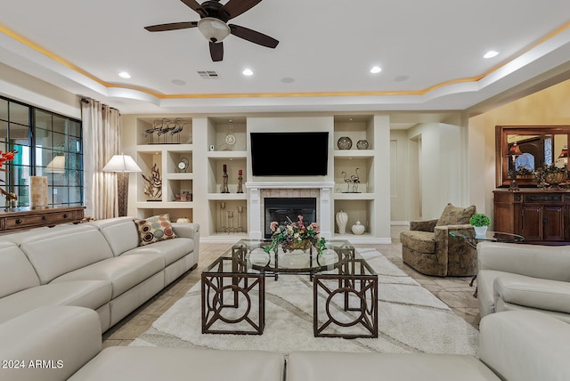 living room featuring a tiled fireplace, built in shelves, a raised ceiling, and ceiling fan