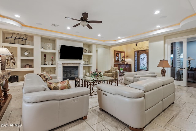 living room featuring ceiling fan, a tray ceiling, a fireplace, and built in shelves