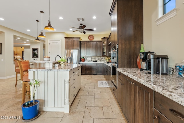 kitchen featuring appliances with stainless steel finishes, backsplash, dark brown cabinets, pendant lighting, and light stone counters