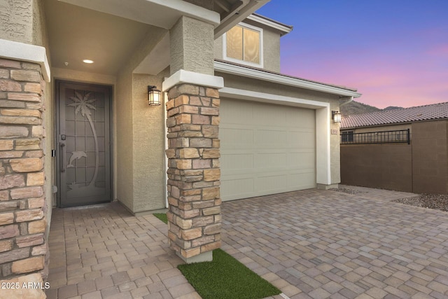 exterior entry at dusk with fence, stucco siding, a garage, stone siding, and decorative driveway