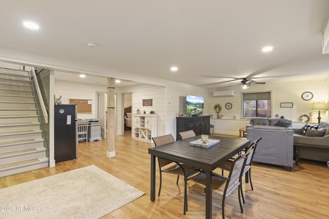 dining area featuring an AC wall unit, stairway, ceiling fan, wood walls, and light wood-type flooring