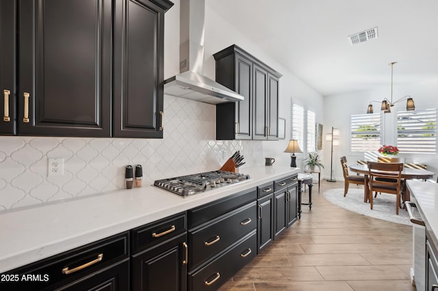 kitchen featuring decorative light fixtures, stainless steel gas stovetop, tasteful backsplash, wall chimney range hood, and light hardwood / wood-style flooring