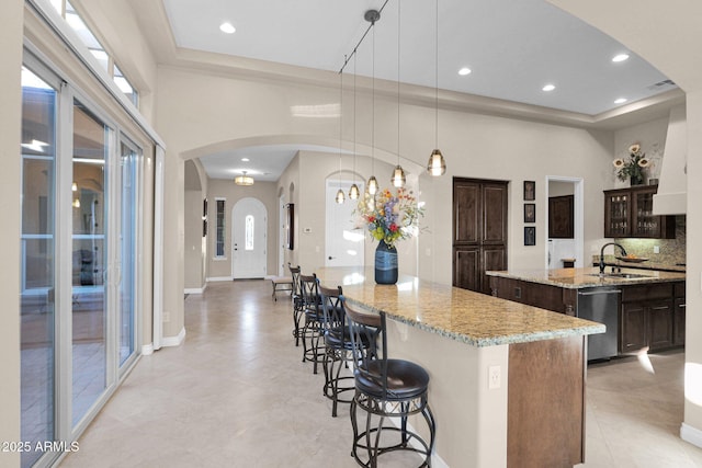 kitchen featuring light stone countertops, stainless steel dishwasher, dark brown cabinets, a kitchen island with sink, and pendant lighting