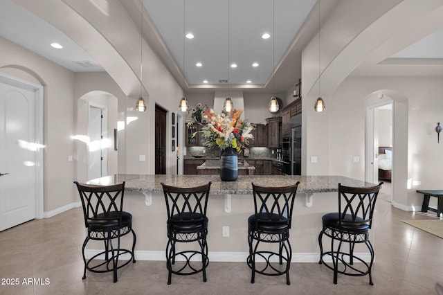 kitchen featuring light stone counters, a breakfast bar, dark brown cabinetry, a tray ceiling, and decorative light fixtures