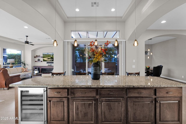 kitchen with light stone countertops, dark brown cabinets, beverage cooler, ceiling fan, and decorative light fixtures