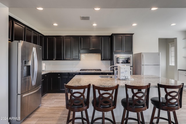 kitchen featuring light hardwood / wood-style flooring, stainless steel appliances, a kitchen breakfast bar, an island with sink, and light stone countertops